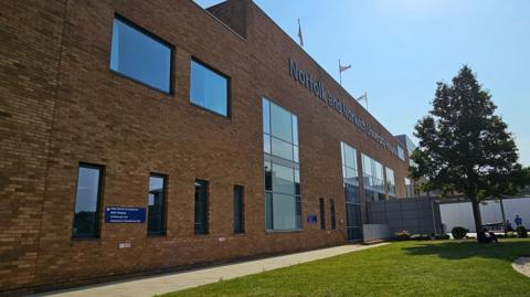 Exterior view of a hospital building. It has brown bricks and a row of tall oblong windows. In the foreground is a pavement, a tree and a grassy area. People can be seen in the distance.
