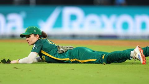 Laura Wolvaardt in green South Africa cricket kit and green cap lies flat out on the turf of the pitch while fielding during the World Cup final against India. She has a look of frustration on her face. In the background is the boundary rope and an advertising hoarding