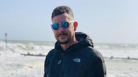 A man with short brown hair and short beard and moustache standing by the sea. He is wearing sunglasses and a padded jacket.