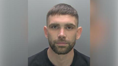 Grant Henry with short dark hair and beard looking at the camera and wearing a black top in a police custody photo