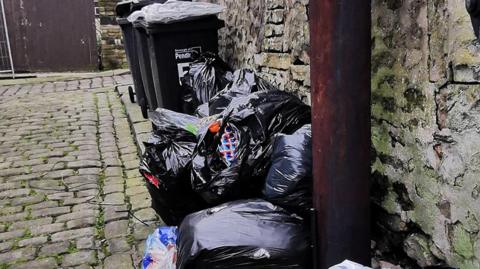 Piles of black bin bags, some ripped open, in a cobbled back alley next to a telegraph pole and three full black wheelie bins