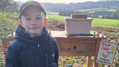 A young boy wears a coat and cap. He is smiling at the camera while standing in front of a wooden desk with signs reading "Sam's Eggs". On the table are two grey plastic boxes, both with a brick on top, and small chick-themed toys.