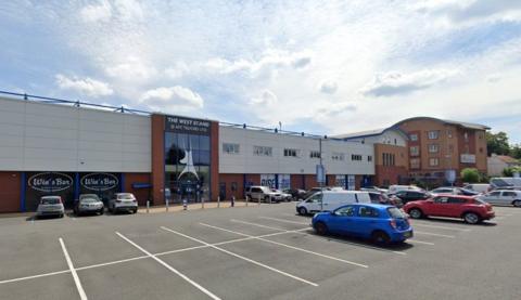 A football stadium car park, with a number of vehicles parked in bays. In front of the car park is a large building with a sign over a glass front entrance that says, "The West Stand". 