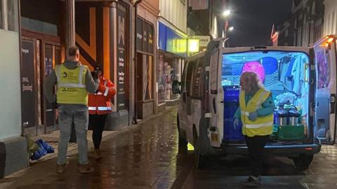 Volunteers deliver food and supplies to a rough sleeper in Hull. They are in a van carrying food, tents and sleeping bags. The man and women are wearing high-vis clothing and gloves.