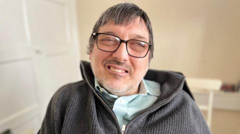 David Cording is pictured sitting down in a room with pale walls. He is wearing a dark grey fleece with a zip over a light blue shirt. He has glasses, light stubble and short dark hair and he is smiling. His eyes are almost closed.