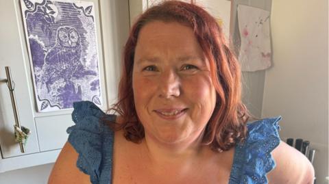 Abbie Barton has shoulder-length red hair and is wearing a blue sleeveless top. She is standing indoors in front of a framed owl print and children's artwork. She is smiling.