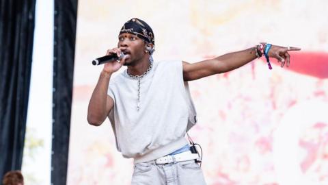 Singer D4vd, wearing a beige vest and black bandana, sings into a microphone.