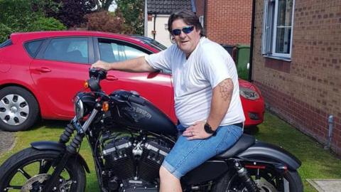Karl Edgeller is sitting on a motorcycle, looking into the camera. He has dark hair and is wearing blue sunglasses, a white t-shirt, and blue denim shorts. He is sitting on a black motorcycle. He has one hand on his hip, one hand on the bike's handles. Behind him, there is a red car. 