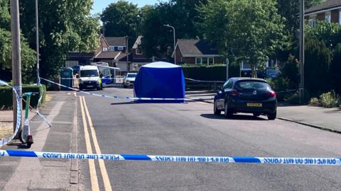 A residential street that has been cordoned off with blue-and-white police tape stretched across the road in several places. In the centre of the scene, a large blue forensic tent has been erected, while police and utility vehicles can be seen further along the road behind the cordon. Parked cars, pavements, streetlights and detached houses line both sides of the street, with mature trees providing a backdrop.