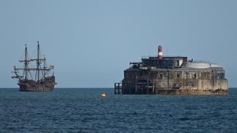 A galleon is on the sea alongside a round fort in the sea of Portsmouth. The replica galleon's sails are stowed and it appears to be anchored.