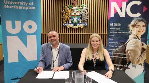 A man in a light blue suit and white open-necked shirt sits alongside a woman with long blonde hair, wearing in a black dress, at a table. Both have pens in their right hands and are signing documents. They are looking at the camera and smiling.