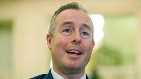 Paul Givan is standing inside the Stormont Building, he is speaking to press. He has short white hair and is wearing a dark suit. 