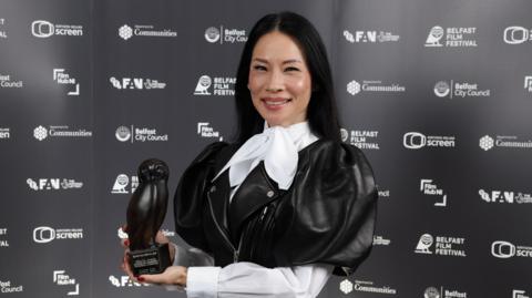 Lucy Liu smiles while posing with her award at the Belfast Film Festival.  She has long, straight, black hair and is wearing a black leather frock coat with puffed sleeves over a white blouse tied with a large bow at her neck.  She is holding an award shaped like an owl. 