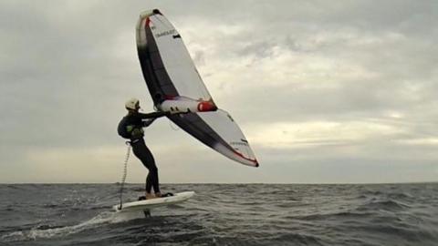 A wingfoiler stands balanced on a hydrofoil board above choppy sea, holding an inflated wing overhead while riding across the water under a cloudy sky.