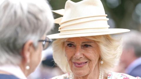 Queen Camilla wears a light coloured hat, pearl earrings and a necklace and a white dress with pink flowers. 