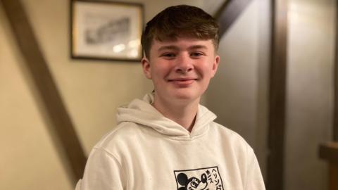 Josh wears a white hoodie with a cartoon logo on it. Behind him is a magnolia-coloured wall with brown timbers forming a 'V' pattern