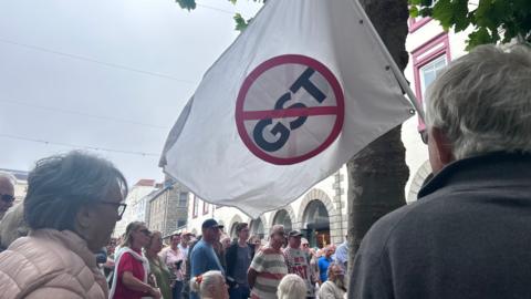 A large flag which says GST in the middle with a red strikethrough going through it. Behind it are people standing some holding anti-GST placards. 