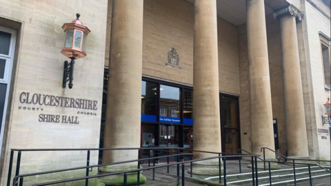 Image shows the outside of Gloucestershire Shire Hall. It is built using limestone and has four pillars outside. 