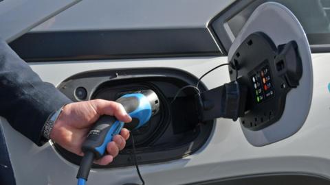 A hand is holding a blue EV charger into the side of a white car. You can see they are wearing a silver watch and dark grey blazer.