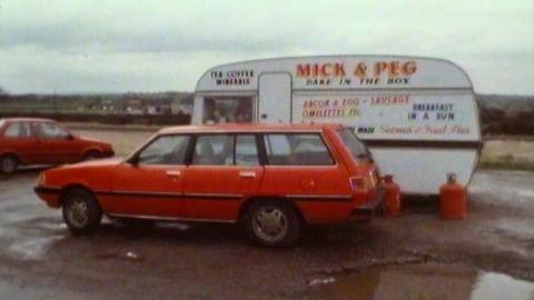 A red estate car parked next to a caravan which sells roadside food to passing drivers.