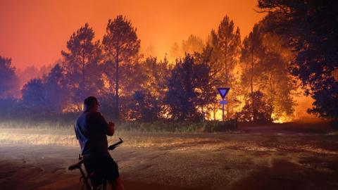 A person leans on a bike in front of a blaze that burns in a forested area
