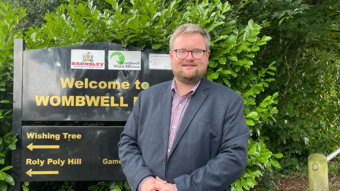 James Higginbottom is wearing a grey suit jacket, dark glasses, and a pale shirt. He is standing in front of a black sign for a park in Barnsley