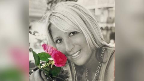 A close up, grey-scale image of Mrs Arter holding some flowers which are in colour - pink petals and a green stem with leaves. She is smiling to the camera and has a necklace on. 