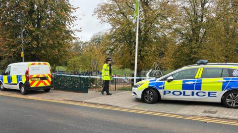 Two police vehicles are parked on a pavement next to a park, which has police tape across the entrance. A police officer stands by the tape and there are autumn trees behind her.