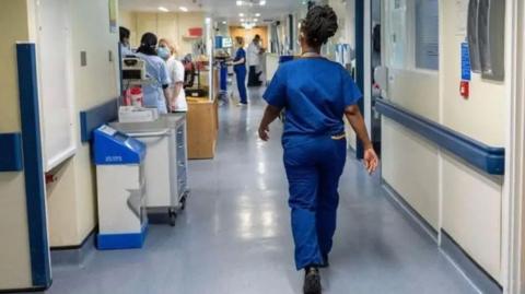 A woman blue wearing scrubs walking through a ward in a hospital. Other nurses and doctors are standing in the background and talking with computers and medical equipment nearby.