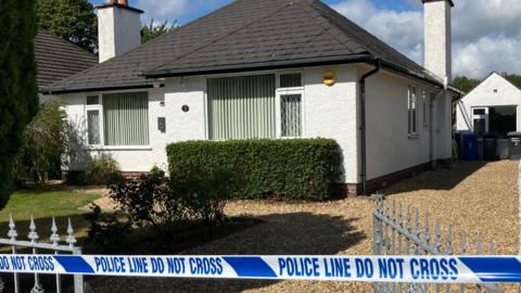 A white bungalow with a black roof with a garden in front of it and a stoned driveway is cordoned off with blue police tape.