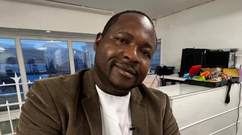 A man sitting in an office looks straight to camera. He is wearing a brown jacket.