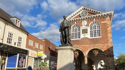 A statue of Sir Robert Peel sits on a stone plinth in front of a prominent brick building with two arched windows and a clock at the top. There are hanging baskets with flowers in them attached to two archways on the lower part of the building.