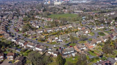 An aerial shot of a low-density suburban neighbourhood with detached and semi-detached houses.
Streets are arranged in curving, cul-de-sac-style layouts, typical of post-war residential planning.
In the background, the landscape transitions into a mix of larger buildings, light industrial units, and open areas.