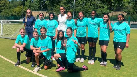 A team of young female football players line up in two rows to pose for a photo with Jill Scott on a green artificial pitch on a sunny day.