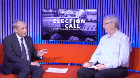 Two men sitting on a red sofa. On the left, the man has short grey hair. He is wearing a dark suit, dark tie and light shirt. On the right, the man has short grey hair and glasses. He is wearing a light blue and white checked shirt and dark trousers. Between them is a TV screen which says 'Election Call Chief Minister's Questions'.