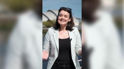 A woman with a brown bob, light blue blazer and sunglasses smiles at the camera. The SEC Armadillo is in the background.