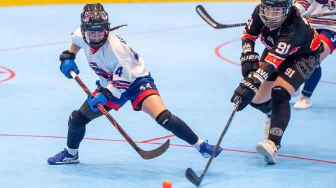 A photo from the Ball Hockey World Championship this year. Iona, wearing a protective cage-helmet on her head and a white jersey adorned by red and blue stripes, is lunging towards the small, orange ball with her stick. But to her left, an opponent in all black kit also dives forward, as they fight for possession.