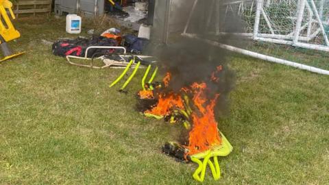 A yellow pop-up football goal on fire laying on the grass outside a storage container. There is other equipment strewn all over the grass and spilling out of the container. There is black smoke coming off the burning goal.