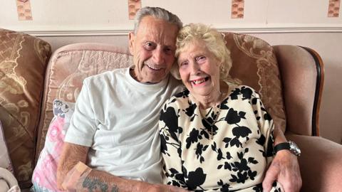 Harry and Rose Brumhead smile at the camera while hugging each other on the sofa with their heads against one another. Harry has grey hair and is wearing a white t-shirt. Rose has blonde, curled hair and is wearing a cream-coloured jumper with black flowers.