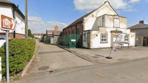 Streetview image of the old village hall - a single storey brick building with a weatherbeaten whitewashed entrance. Two signs point down a side road to the mining museum.