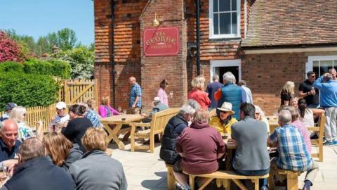 Dozens of people sat on benches in a beer garden outside a pub on a sunny day