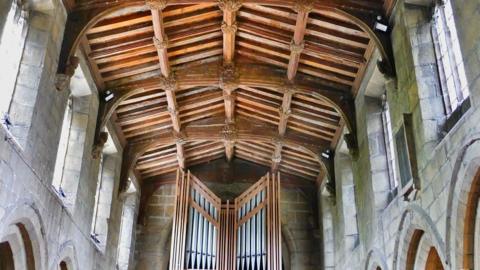 Interior of an exposed wooden church roof, below are organ pipes surrounded by stone walls