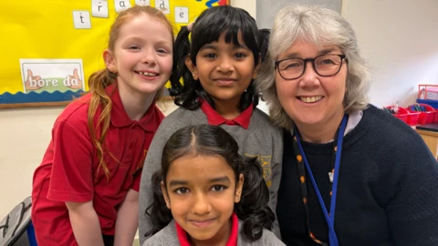 Tair merch fach mewn gwisg ysgol a'u hathrawes yn gwenu ar ycamera 