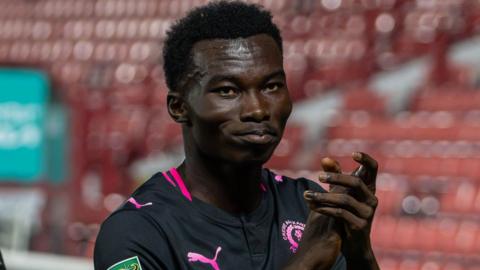 Crewe Alexandra forward Joel Ewusi applaudes the fans after becoming the club's youngest ever appearance maker and scorer. He is wearing a black shirt with pink edging and writing on it.