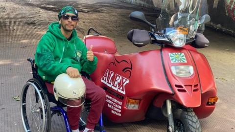 David Cappelletti sat in his wheelchair wearing a green jumper with his thumbs up. He is sitting alongside his red bike that was stolen