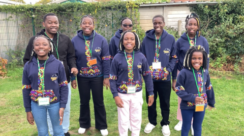 Members of the choir stand outside and are all smiling at the camera. With the exception of one man, they are all wearing navy hoodies with a colourful pattern down the middle. They are also wearing green lanyards. The outdoor setting they are in has grass on the floor and a fence behind them.