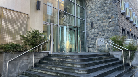 Frontage of Guernsey royal courts. Steps leading up to a glass entrance with a crest above it.
