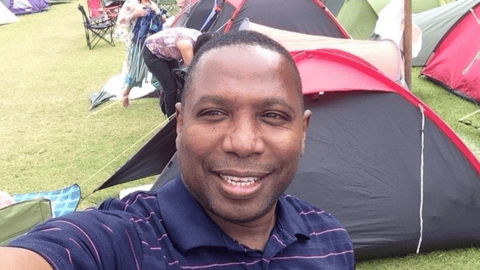 A man in a navy polo shirt taking a selfie. He is standing in front of a line of tents.