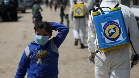 An internally displaced Syrian girl wears a face mask as members of the Syrian Civil defence sanitize the Bab Al-Nour camp