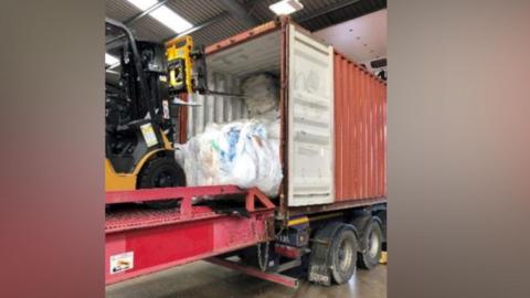 A yellow forklift truck is on a red lift attached to the back of a lorry with a shipping container on the back. The forklift has been moving white sacks into the back of the container.